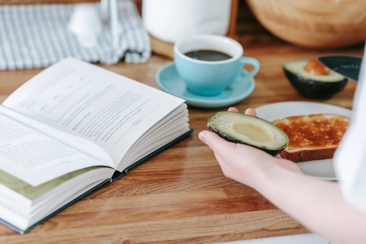 An open book beside a cup of coffee and breakfast on a kitchen table